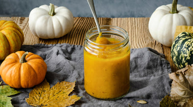 Jar of mashed pumpkin for dogs with a spoon inside. The jar is surrounded by different coloured pumpkins.